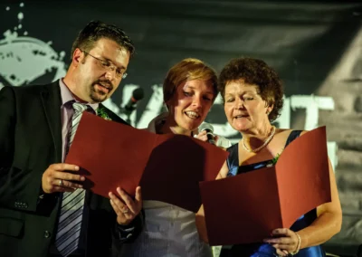 Drei Personen lesen lächelnd von roten Mappen ab, eine Frau hält ein Mikrofon, Hintergrund dunkel.