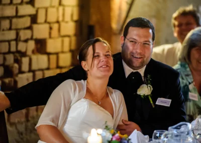 Ein lachendes Paar sitzt dicht beieinander, beide in festlicher Kleidung, umgeben von Kerzenschein und Gästen.