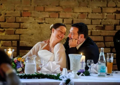 Ein Paar sitzt lachend an einer geschmückten Tafel mit Kerzen und Getränken vor einer Backsteinwand.