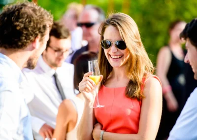 Eine Frau im korallenfarbenen Kleid mit Sonnenbrille lächelt, hält ein Glas Sekt, umgeben von entspannt plaudernden Gästen.