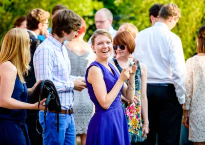 Eine lachende Frau in einem lilafarbenen Kleid hält eine Kamera, umgeben von elegant gekleideten Gästen im Freien.