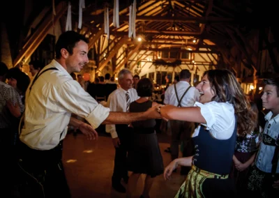 Ein Paar in traditioneller Tracht tanzt lachend in einem festlich beleuchteten Saal, umgeben von weiteren Gästen.