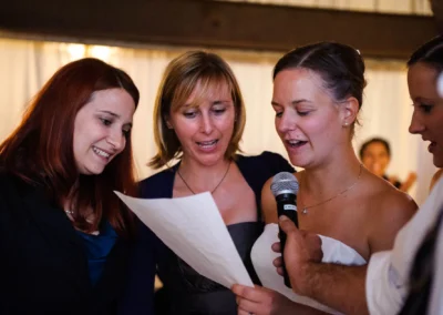 Vier Personen stehen zusammen, eine Frau im weißen Kleid hält ein Mikrofon und ein Blatt, sie schauen darauf und wirken konzentriert.