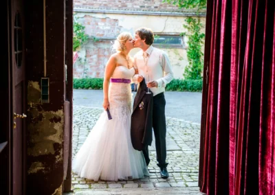 Ein Brautpaar küsst sich im Freien. Die Braut trägt ein weißes Kleid mit lila Band, der Bräutigam hält ein Glas Sekt.