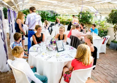 Gäste sitzen an weiß gedeckten Tischen mit Blumenschmuck, unter einer überdachten Terrasse, in einer lebhaften Unterhaltung.