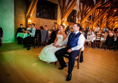 Ein Brautpaar sitzt lächelnd auf Stühlen, Gäste in festlicher Kleidung schauen zu, beleuchteter Raum mit Holzbalken.