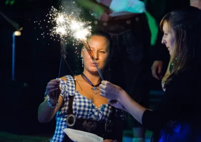 Zwei Frauen in Tracht halten Wunderkerzen, eine lächelt. Hintergrund dunkel, Partystimmung.
