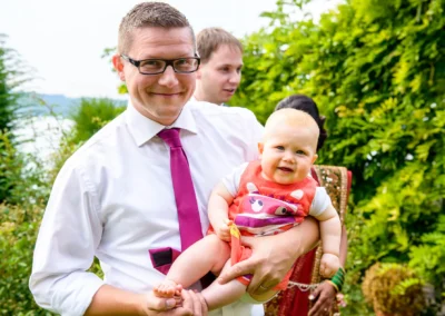 Mann haelt lachendes Baby bei Hochzeit im Garten des Seehotel Leoni am Starnberger See. Hochzeitsreportage.