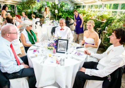 Gäste sitzen um einen festlich gedeckten Tisch mit Blumen und Fotos. Die Braut lächelt, das Brautpaar ist in eleganter Kleidung.