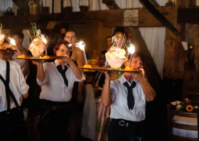 Bedienungen tragen Tabletts mit brennenden Wunderkerzen und Eisbechern durch einen festlich geschmückten Raum.