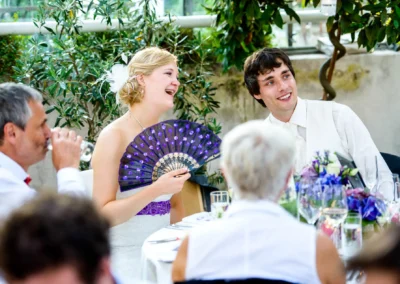 Braut hält einen Fächer, beide lachen am Tisch, geschmückt mit Blumen, Gäste im Hintergrund, festliche Atmosphäre.