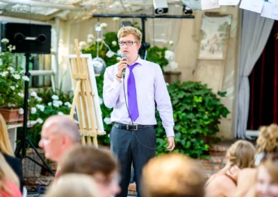 Ein Mann in Hemd und Krawatte hält eine Rede, Mikrofon in der Hand, Pflanzen im Hintergrund, mehrere Personen im Vordergrund.