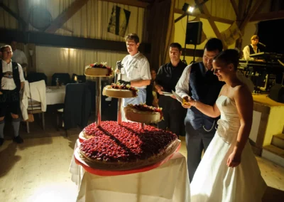 Brautpaar schneidet gemeinsam eine dreistöckige Hochzeitstorte an. Im Hintergrund stehen Gäste, einer in traditioneller Kleidung.