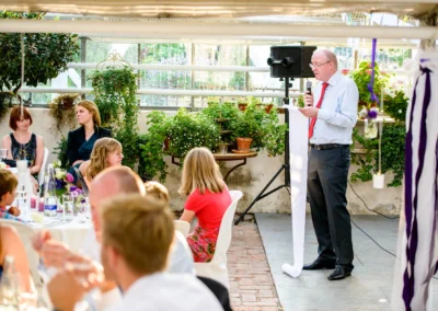 Ein Mann hält eine Rede mit Mikrofon, Gäste sitzen an gedeckten Tischen im Gewächshaus, Pflanzen im Hintergrund.