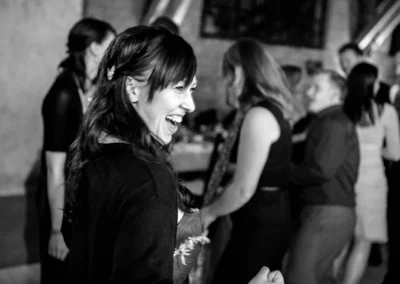 Eine lachende Frau in einem festlichen Raum, umgeben von mehreren tanzenden Menschen in eleganter Kleidung.