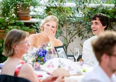 Ein Brautpaar lacht fröhlich am Tisch; Blumendekoration und Sektgläser sind sichtbar. Eine Frau mit Fächer ist im Vordergrund.