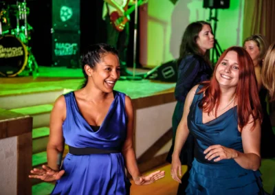 Zwei lachende Frauen in blauen Kleidern tanzen vor einer Bühne mit Instrumenten und grüner Beleuchtung.