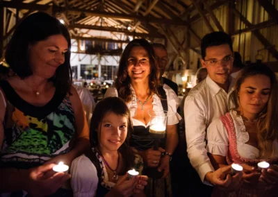 Menschen in festlicher Kleidung halten Kerzen in einem warm beleuchteten Raum mit Holzbalken. Sie lächeln und wirken entspannt.