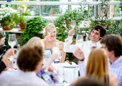 Das Brautpaar stößt mit Gästen an, lachend an einem gedeckten Tisch in einem grünen, blumenreichen Gewächshaus.
