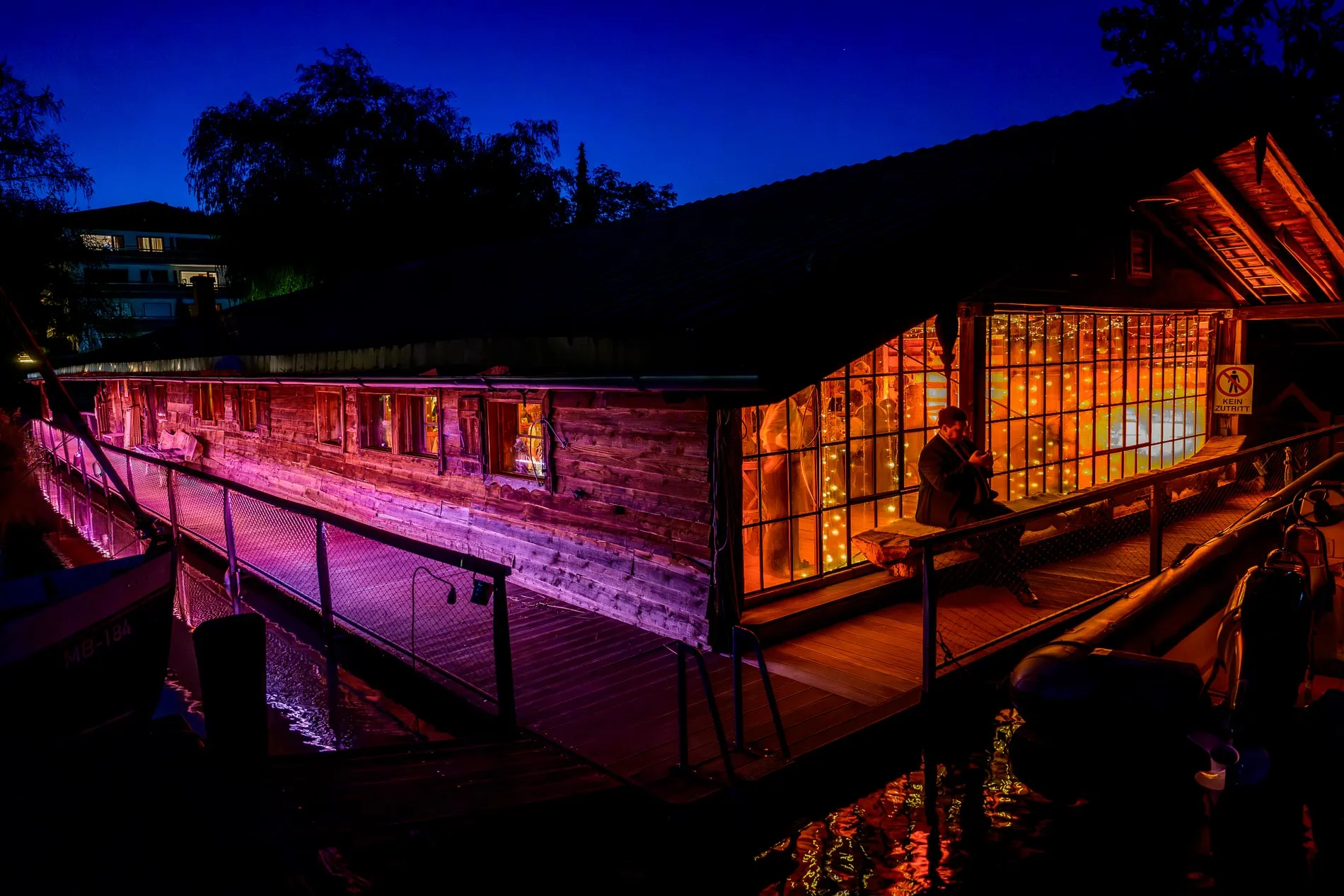 hochzeitsfotografie-feier-tegernsee Altes Gebäude am Wasser, beleuchtet in violett und orange, Innenraum voller Lichterketten, Person sitzt außen auf der Bank.