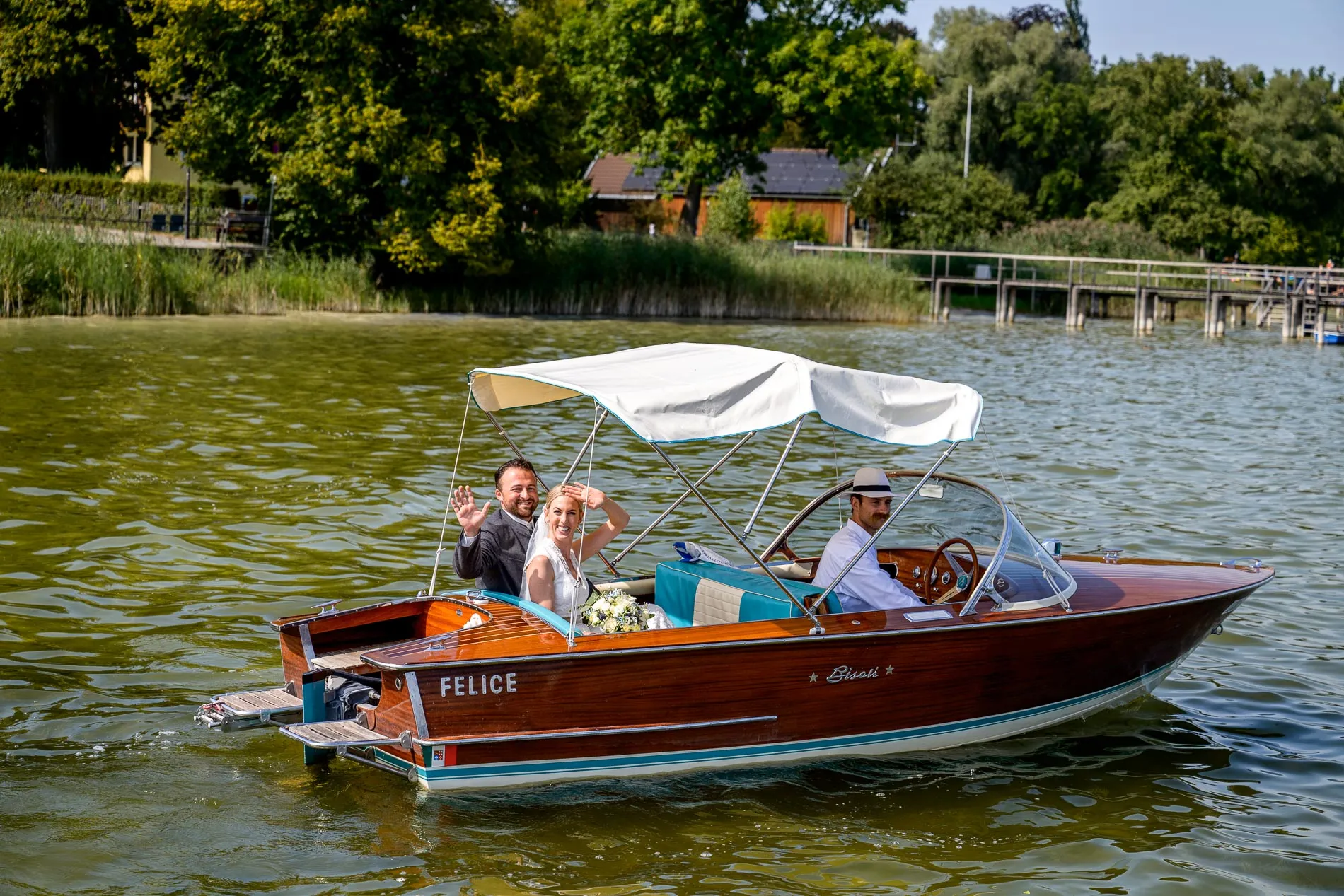 hochzeitsreportage-ammersee Ein Brautpaar sitzt lächelnd in einem kleinen Holzboot auf einem See, ein Mann mit Hut steuert es, grüne Ufervegetation im Hintergrund.