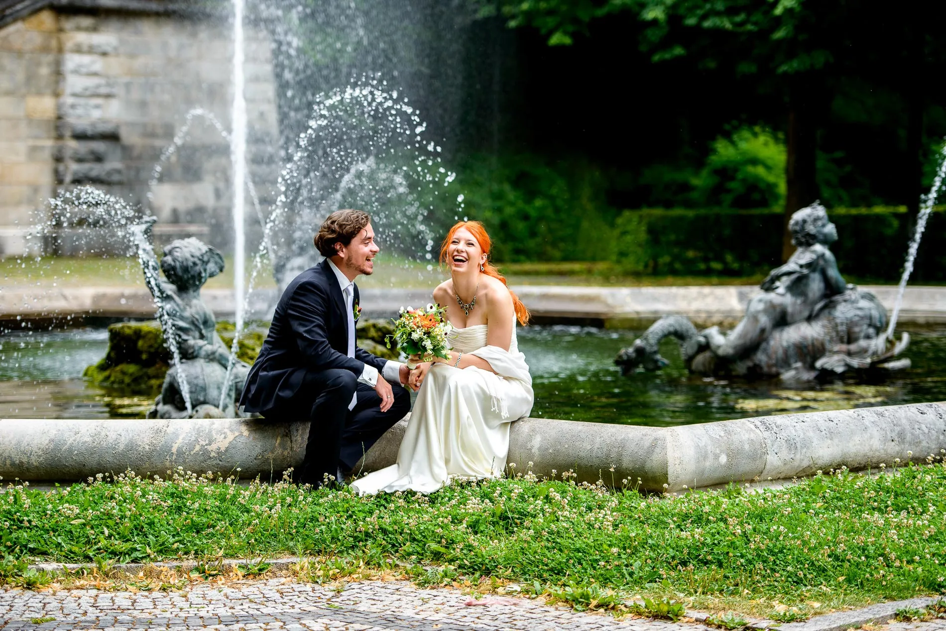 muenchen-hochzeit Hochzeitsfoto eines Brautpaars das lachend lachend am Brunnen unterhalb des Friedensengels in München sitzt